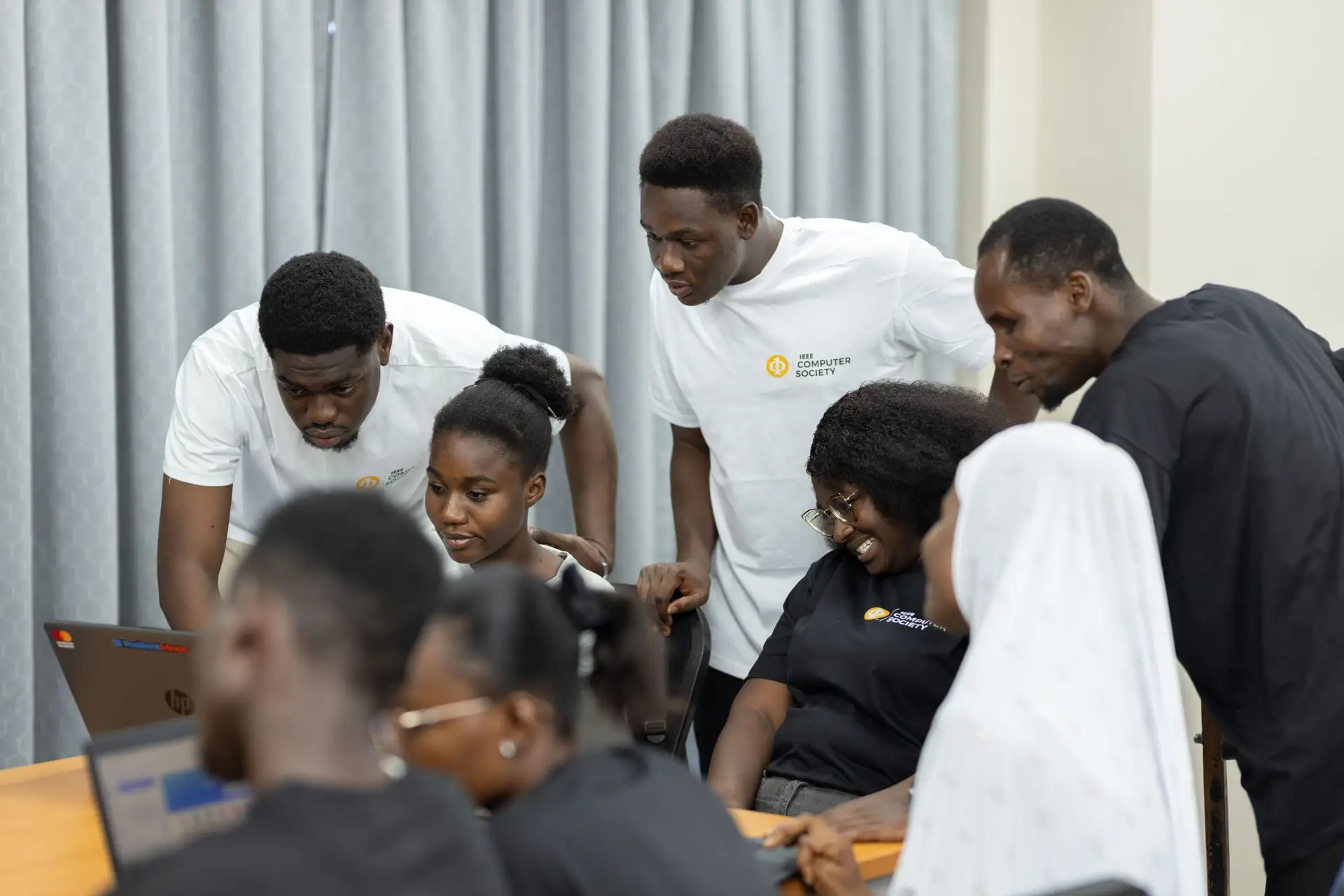 A group of young adults collaborates around laptops in a classroom, with some wearing IEEE Computer Society shirts.
