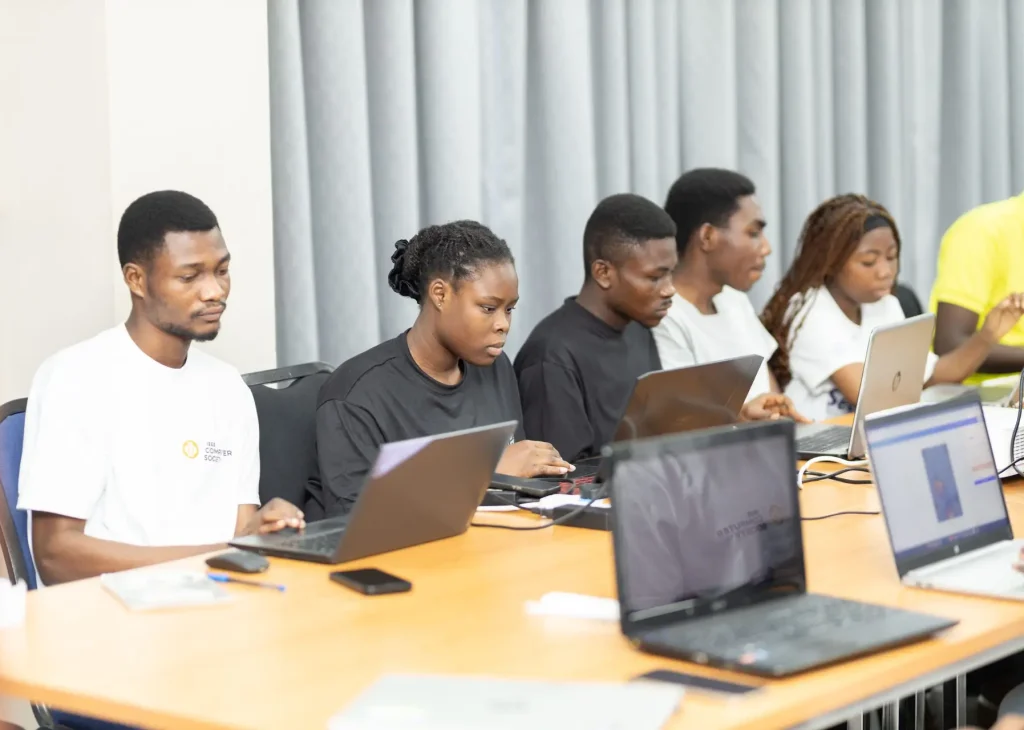A group of young adults sit at a long table focused on laptops during a training session.