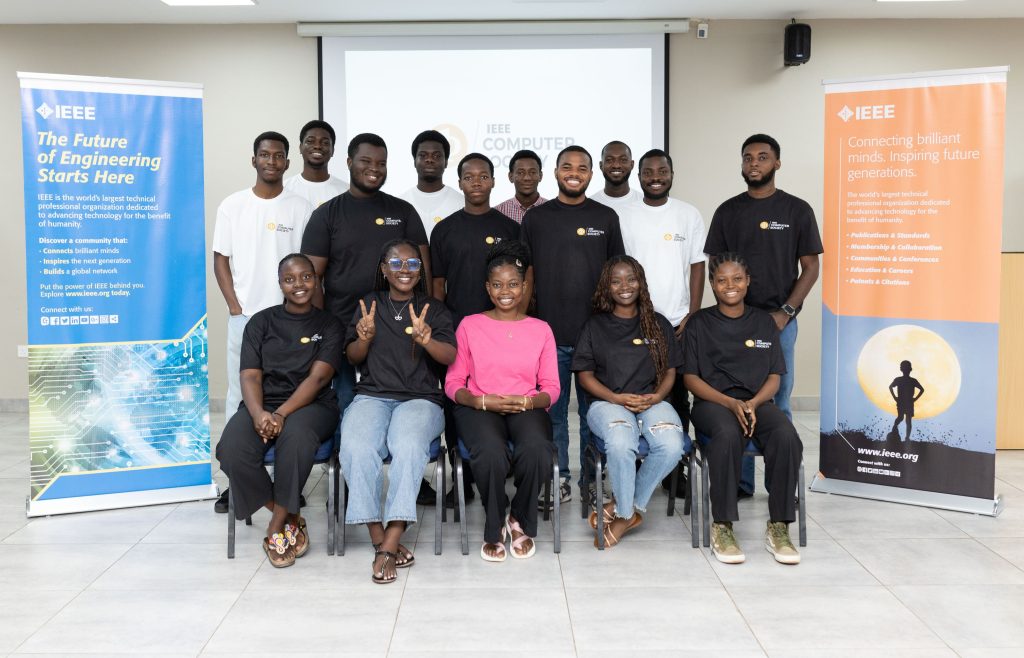 A group of 15 young adults pose for a photo between IEEE banners at an engineering or computer society event.