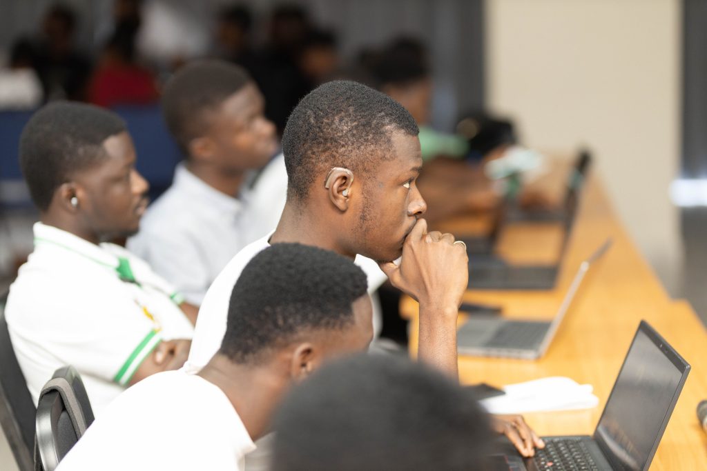 A group of young Black men sitting at a long table with laptops in a classroom setting, with one man in the foreground wearing a hearing aid and looking thoughtfully ahead.