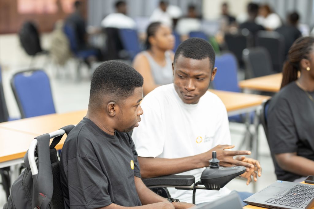 Two young men in a classroom setting are engaged in conversation, with one man sitting in a motorized wheelchair.