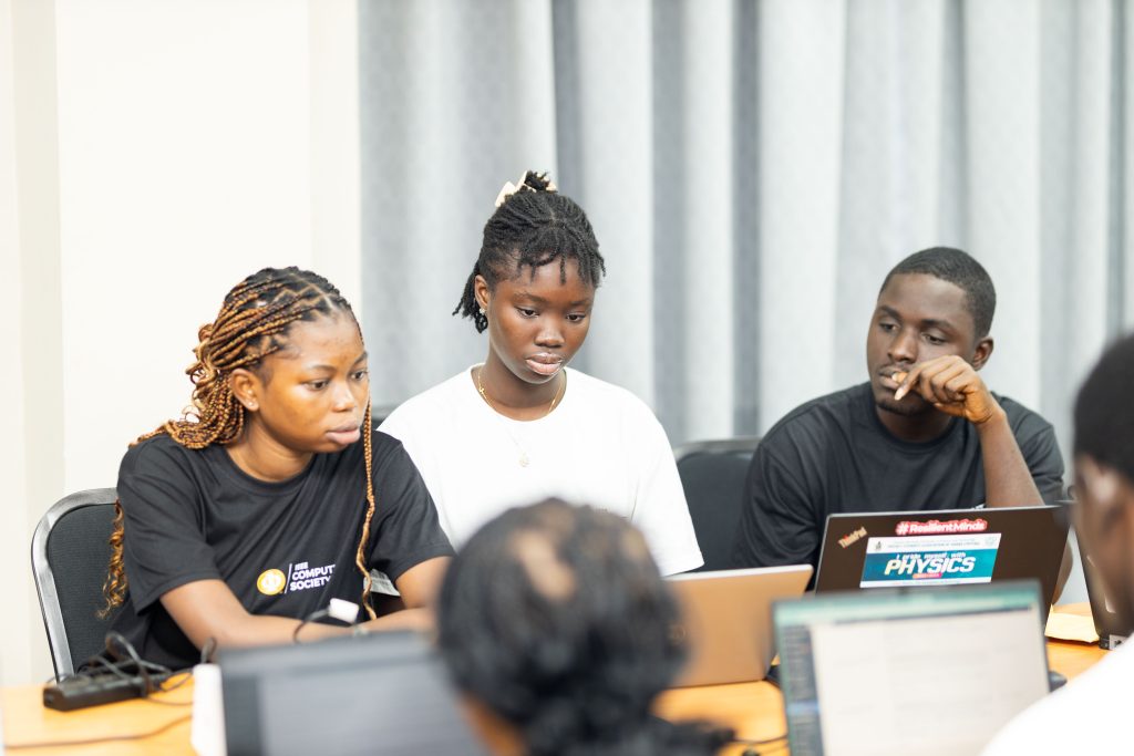 Three young Black students sit at a table with laptops, focused on a screen during a collaborative session.