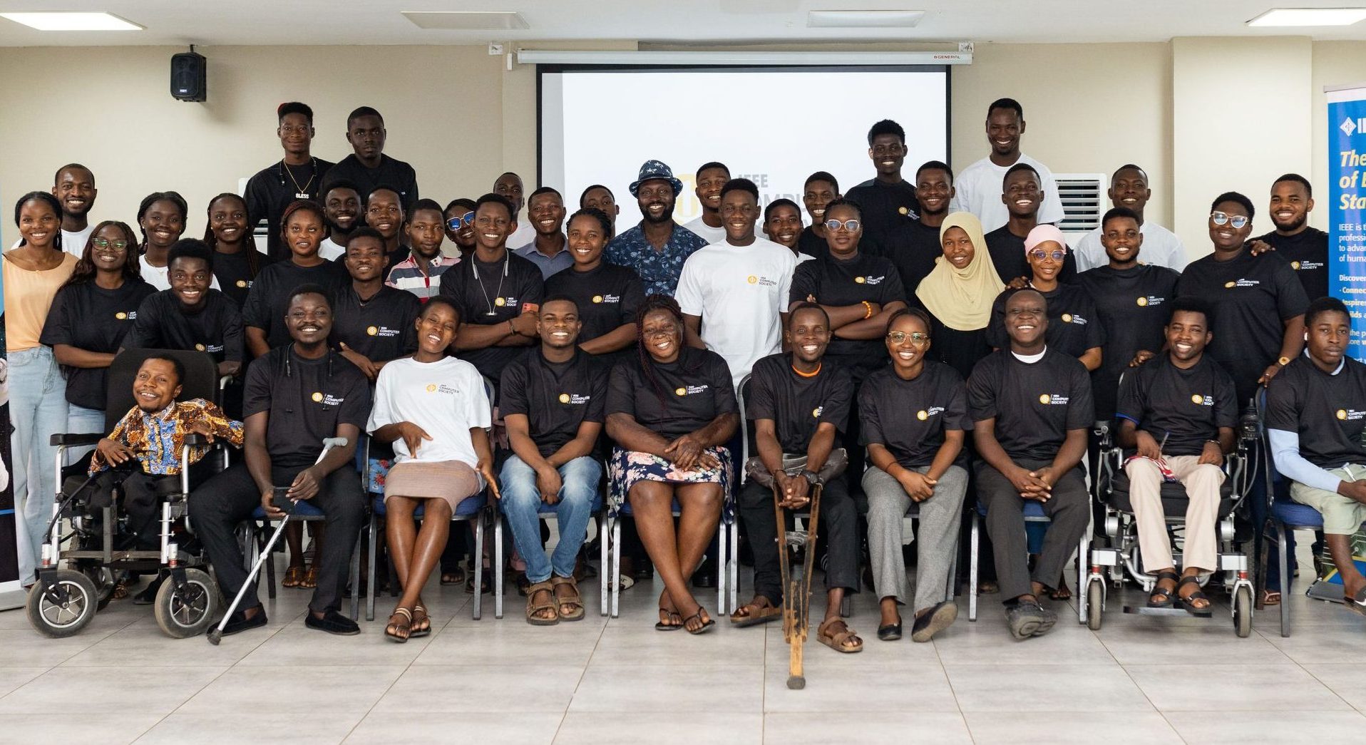 A large diverse group of young people, including several wheelchair users, pose together for a photo in a conference room flanked by IEEE banners.