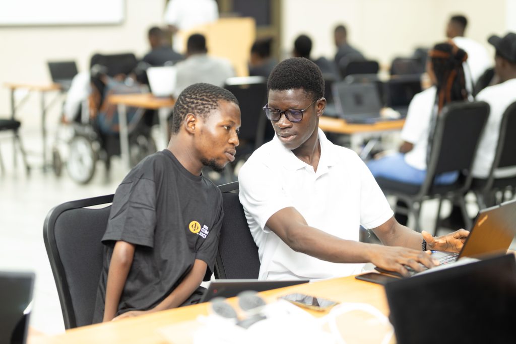 Two young men collaborate on a laptop during a computer training session, with other participants working in the background.