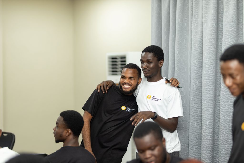 Two young men wearing IEEE Computer Society shirts smile together with their arms around each other during an indoor gathering.